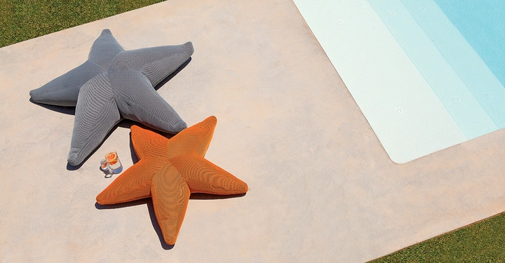 Zwei sternförmige Kissen liegen auf einer Betonfläche neben einem Swimmingpool ein großes graues und ein etwas kleineres orangenes; daneben steht ein Glas mit Getränk und eine Sonnenbrille auf dem Boden.