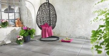 Hängesessel hängt in einem hellen Raum mit grauen Fliesen und weißer Wanddeko Sitzkissen und Decke darin Ein Buddha aus Holz und Pflanzen auf der Fensterbank Blumen und Teeservice auf Boden nearby