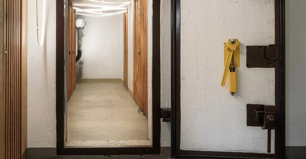 Blick in einen modernen Schutzraum mit massiver Stahltür und Gang zu Holzverschlägen. Hausbunker im Keller eines Einfamilienhauses