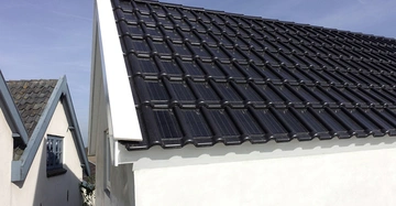Schwarze Solardachziegel bedecken das Satteldach eines weißen Hauses im Außenbereich An den Seiten sind zwei weitere Häuser mit traditionellen Dachziegeln sichtbar darunter ein blauer Himmel
