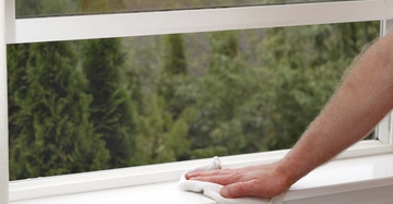 Eine Hand wischt mit einem weißen Tuch eine Fensterbank sauber in einem Raum mit Blick auf Bäume durch das offene Fenster.