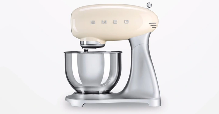 Küchenmaschine mit cremefarbenem Kopf und Edelstahlrührschüssel steht in einem minimalistisch weißen Raum. Text auf dem Kopf SMEG