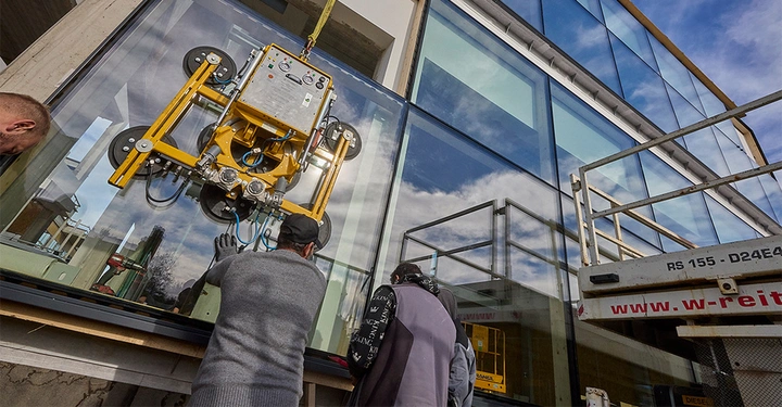 Arbeiter installieren mit einem gelben Vakuumheber eine große Glasscheibe an einem modernen Gebäude mit Spiegelverglasung in einem städtischen Umfeld bei bewölktem Himmel Text: w-reitz.com RS 155 D24E4