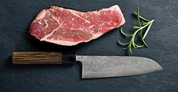 Rohes Steak liegt neben einem Küchenmesser und einem Rosmarinzweig auf einer dunklen Schieferplatte.