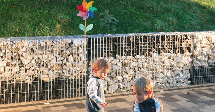 Zwei Kinder spielen draußen Einer fährt ein rotes Dreirad ein anderer steht dahinter Im Hintergrund ein grüner Hügel mit einem farbigen Windrad und einem steinernen Zaun.