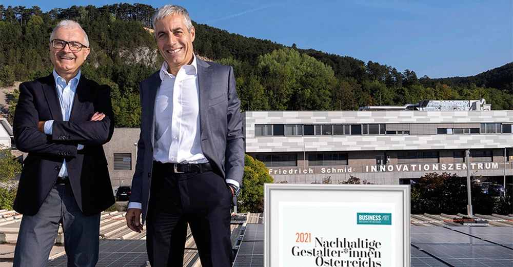 Zwei Männer posieren lächelnd in Anzügen. Im Hintergrund befindet sich das Friedrich Schmid Innovationszentrum vor einer bewaldeten Landschaft. Text: 2021 Nachhaltige Gestalter*innen Österreichs BUSINESS ART