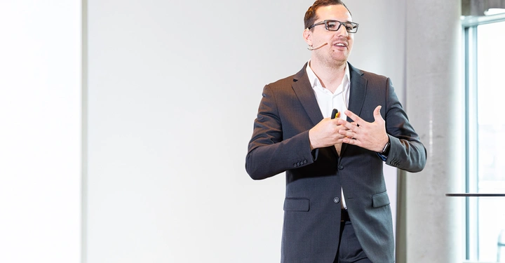 Galerie-Bild 4: Ein Mann im Anzug spricht in einem modernen hellen Raum steht vor einem neutralen Hintergrund trägt ein Mikrofon und nimmt eine gestische Position ein während er ein Publikum anspricht.