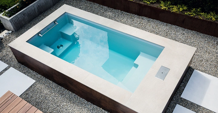 Rechteckiges Schwimmbecken mit klarem Wasser im Garten umgeben von Kieselsteinen und Pflastersteinen mit einem kleinen bepflanzten Bereich im Hintergrund