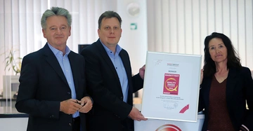 Drei Personen halten eine gerahmte Urkunde in einem modernen Büro. Text auf der Urkunde: MARKTINSTITUT MARKTTEST QUALITY AWARD 2021 MARKET INSTITUT BERKER Für die stärkste Weiterempfehlung in der Kategorie SCHALTER & MELDESYSTEME