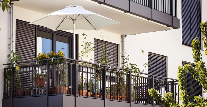 Galerie-Bild 1: Moderner Balkon mit schwarzem Geländer, vielen Pflanzen in terrakottafarbenen Töpfen und einem aufgespannten weißen Sonnenschirm