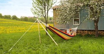 Eine Person liegt in einer bunten Hängematte und liest ein Buch neben einem blühenden Baum und einem Holzhaus auf einer grünen Wiese mit gelben Blumen im Hintergrund