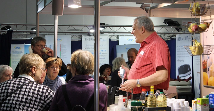 Galerie-Bild 10: Ein Mann im roten Hemd erklärt einer Gruppe älterer Menschen in einer Messehalle die Verwendung eines Küchengeräts. Im Hintergrund befinden sich Werbebanner und diverse Produkte auf dem Tisch.