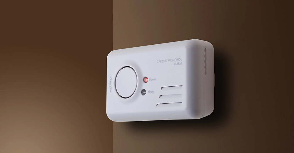 Ein weißer Kohlenmonoxidmelder mit Leuchten montiert an einer braunen Wand. Auf dem Gerät sind die Worte CARBON MONOXIDE ALARM sowie TEST RESET zu lesen.