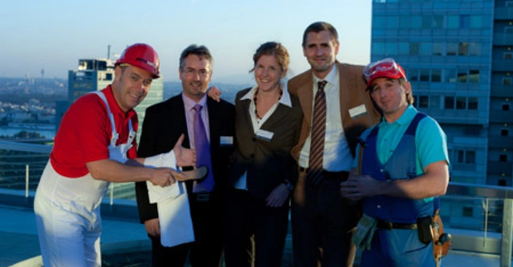 Galerie-Bild 2: Fünf Personen stehen auf einem Dach einer Stadt im Hintergrund Zwei tragen Arbeitsoveralls und Helme drei tragen formelle Kleidung Sie lächeln und posieren für die Kamera.