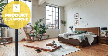 Bett mit Decken und Kissen im Schlafzimmer mit großen Fenstern und Pflanzen umgeben. Es gibt eine gelbe Blase oben links mit dem Text: 7 PRODUKT DER WOCHE.