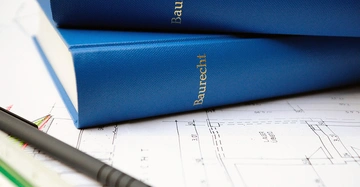2 blaue dicke Gesetzbücher mit Schriftzug BAURECHT in silber liegen auf einem Hausplan am Tisch. Kugelschreiber und Lineal darauf.