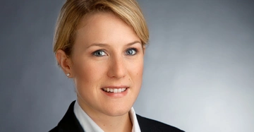 Frau lächelt, trägt einen schwarzen Blazer und eine weiße Bluse, vor einem neutralen grauen Hintergrund.