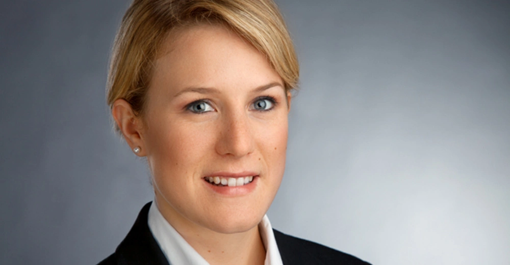 Frau lächelt, trägt einen schwarzen Blazer und eine weiße Bluse, vor einem neutralen grauen Hintergrund.