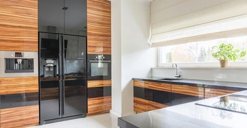 Großer schwarzer Kühlschrank eingebaut in eine Schrankwand aus Holz Küchenarbeitsplatte aus schwarzem Stein darunter Spüle mit Wasserhahn rechts großes Fenster mit weißem Rollo und grüne Pflanze im Topf