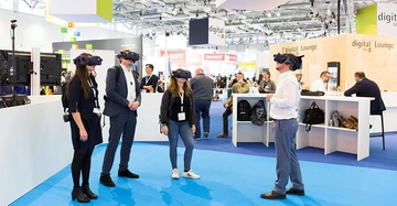 Vier Personen mit VR-Headsets stehen in einem Messebereich während eine Person spricht andere beobachten Im Hintergrund befinden sich Informationsstände und weitere Besucher. Schildern: digital BAU sowie Digital Lounge.