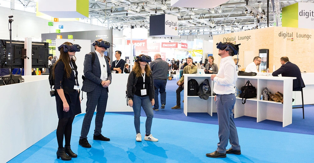 Vier Personen mit VR-Headsets stehen in einem Messebereich während eine Person spricht andere beobachten Im Hintergrund befinden sich Informationsstände und weitere Besucher. Schildern: digital BAU sowie Digital Lounge.