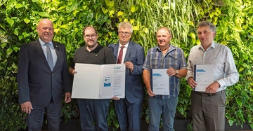 Fünf Männer in Businesskleidung halten Zertifikate in einer Indoor-Pflanzenwand stehend Hände schütteln und lächeln Texte: "EcoZert 2023" "CERTIFICATION BILANZANALYSE 2023"