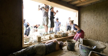 Gruppe von Studierenden bei der Arbeit in und vor einem Gebäude. Lehmbau mit Holz, es wird verputzt, verspachtelt und gestampft.