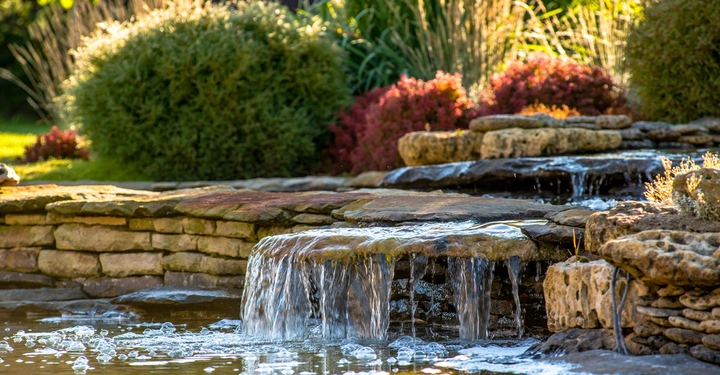 Wasser fließt über gestapelte Natursteine und bildet einen kleinen Wasserfall im Garten. Im Hintergrund sind verschiedene Büsche und Gräser zu sehen.