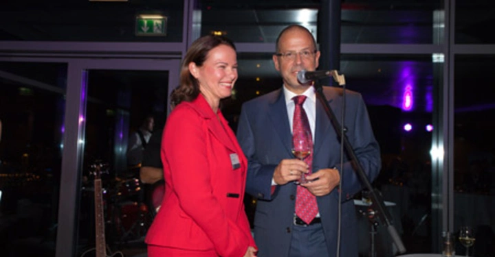 Galerie-Bild 78: Ein Mann im Anzug hält ein Getränk und spricht an einem Mikrofon neben einer Frau im roten Anzug die lächelnd steht in einer eleganten Innenraumumgebung mit Gitarren im Hintergrund