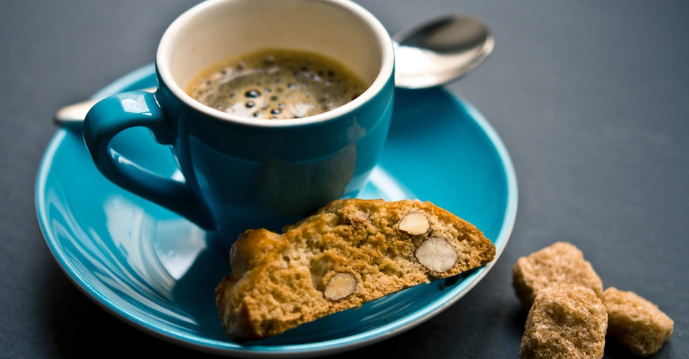 Eine blaue Tasse mit Kaffee steht auf einer blauen Untertasse, auf der auch ein Mandelkeks liegt. Daneben befinden sich Zuckerwürfel auf einer dunklen Oberfläche.