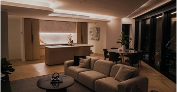 Eleganter, offen gestalteter Wohn- und Essbereich mit moderner Kücheninsel, gemütlichem Sofa, großflächigen Fenstern und stimmungsvoller indirekter Beleuchtung in einer hochwertigen Stadtwohnung bei Nacht.