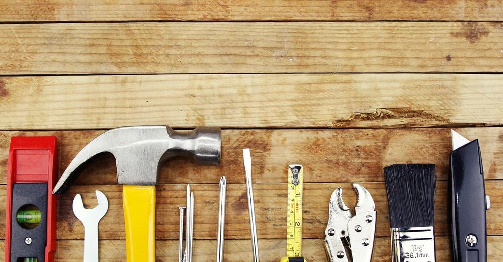 Werkzeuge liegen nebeneinander auf einem hölzernen Untergrund Hammer Wasserwaage Schraubenschlüssel Nägel Schraubendreher Maßband Zange Pinsel Teppichmesser.