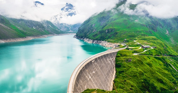 Damm sperrt smaragdgrünen Stausee ab umgeben von üppig grünen Bergen mit verschneiten Gipfeln unter bewölktem Himmel.