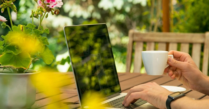 Terassentisch aus Holz mit Kaffeetasse und Bank im Hintergrund. Junge Frau sitzt an Laptop und arbeitet.