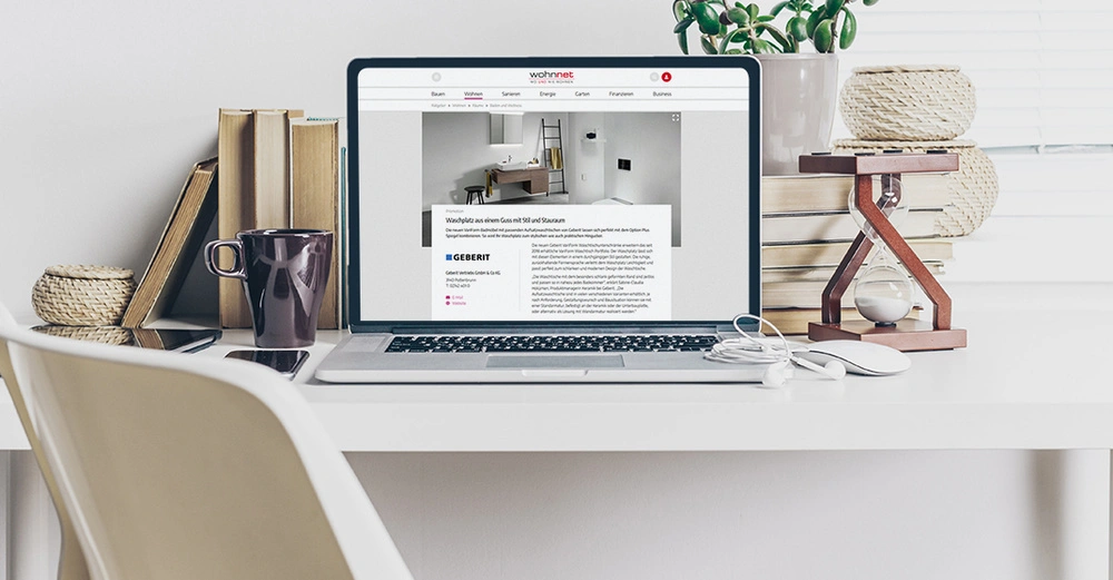 Ein Laptop zeigt eine Website auf einem Schreibtisch neben Büchern einer Pflanze einer Sanduhr und einer Tasse Kaffee. Auf der Website steht oben wohnnet und zeigt einen Artikel über Waschplätze.