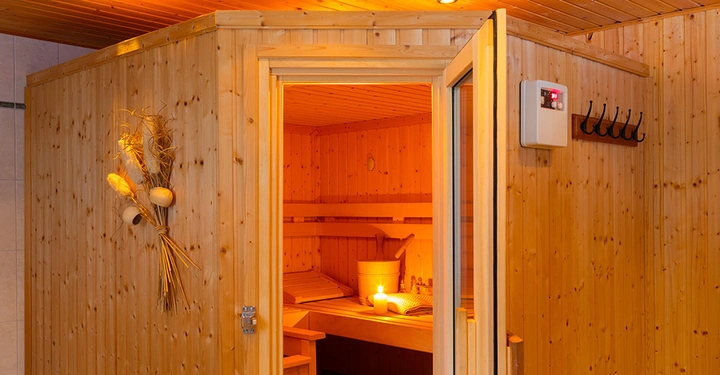 Finnische Heimsauna aus hellem Holz mit geöffneter Tür und Blick auf die Sitzbank und das Kühlwasser im Inneren.