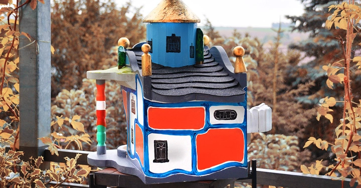 Galerie-Bild 3: Bunt bemaltes Vogelhäuschen steht auf Metallständer in einem herbstlichen Garten umgeben von Bäumen und Sträuchern