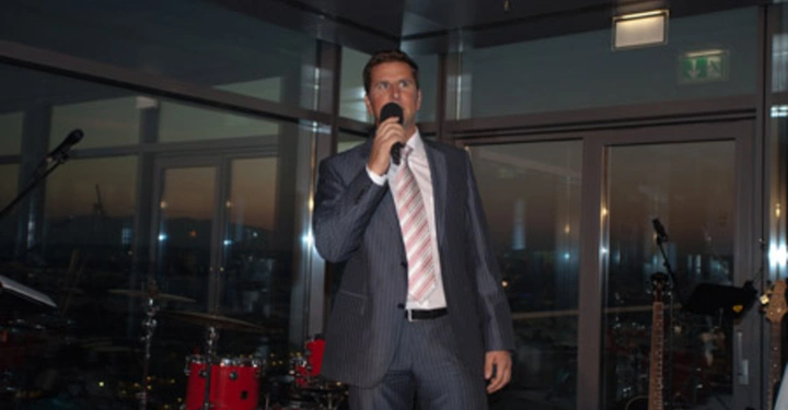 Galerie-Bild 62: Mann im Anzug spricht in Mikrofon steht in einem Raum mit Glaswänden im Hintergrund sind Musikinstrumente wie ein Schlagzeug und Gitarren erkennbar Beleuchtung ist gedämpft und es ist abends.