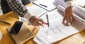 Person zeichnet mit Lineal und Stift auf großen Bauplänen an einem Holztisch; umgeben von Laptop Tastatur Notizbuch Handy und Stiften in einem Büro oder Arbeitsraum