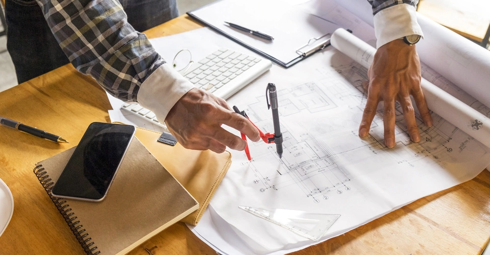 Person zeichnet mit Lineal und Stift auf großen Bauplänen an einem Holztisch; umgeben von Laptop Tastatur Notizbuch Handy und Stiften in einem Büro oder Arbeitsraum