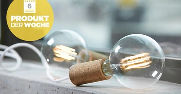 Eine beleuchtete Glühbirne mit Steckdose ruht auf einer Betonfläche vor einem Fenster mit unscharfem Hintergrund. Text: 6 PRODUKT DER WOCHE