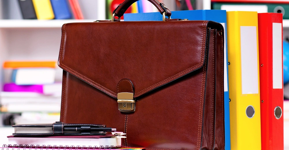 Eine braune Ledertasche steht auf einem Schreibtisch neben bunten Ordnern. Notizbücher und Stifte liegen vor der Tasche. Im Hintergrund befindet sich ein Regal mit weiteren Ordnern und Bürozubehör.