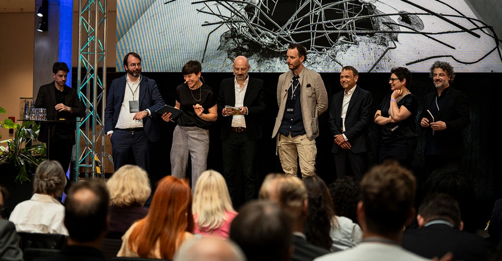 Acht Personen, davon zwei Frauen, stehen lachend und leger gekleidet auf einer dunklen Bühne. Im Hintergrund große grüne Pflanzen. Ausgelassene Stimmung.