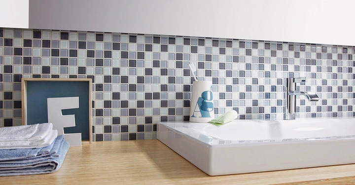 Galerie-Bild 8: Ein weißes Waschbecken auf einer Holzoberfläche mit einem Zahnbürstenbecher und einem Seifenstück darauf vor einem Mosaikhintergrund aus blauen und grauen Fliesen. Links gefaltete Handtücher und ein gerahmtes Bild mit einem großen Buchstaben E