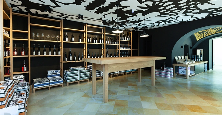 Galerie-Bild 8: Holztisch steht still in einem Raum mit Weinflaschen auf Regalen an der Wand und Kartons auf dem Boden unter schwarz-weiß gemusterten Deckenlampen in einem modernen Geschäft.