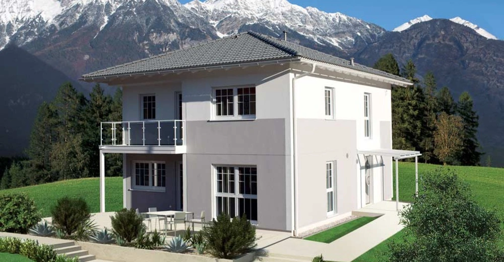 Ein zweistöckiges Haus steht auf einer grünen Wiese im Vordergrund schneebedeckte Berge im Hintergrund umgeben von dichten Wäldern der Himmel ist klar und blau.