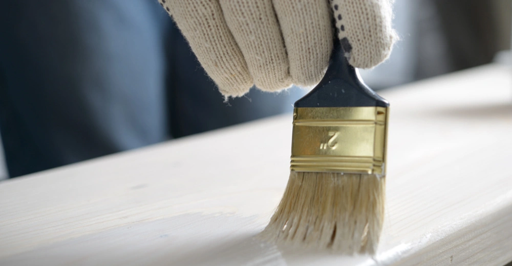 Hand in Strickhandschuh hält Pinsel streicht weiße Farbe auf Holzoberfläche im Innenraum.