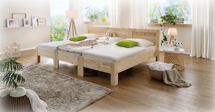 Ein Holz-Doppelbett mit weißen Bettlaken und grünen Kissen steht in einem hellen Schlafzimmer mit Holzfußboden. Im Hintergrund befinden sich große Fenster, Pflanzen und ein Teppich.
