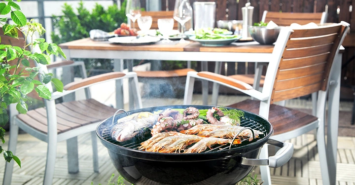 Runder Grill voller Meeresfrüchte und Gemüse grillt Rauch steigt auf Im Hintergrund ein gedeckter Tisch mit Holzstühlen steht auf einer Terrasse mit grünen Pflanzen.