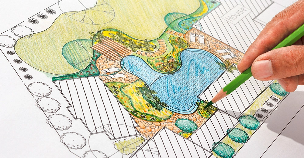 Eine Hand färbt mit einem grünen Bleistift einen Landschaftsplan aus mit einem zentralen Schwimmbecken umgeben von Pflanzen und Gehwegen. Oberes Feld markiert als House